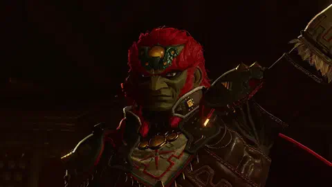 Close-up of Ganondorf's face in the dark: red hair, green skin, amber eyes, a green-and-gold jewelled piece on his forehead, dark armor with red Gerudo patterning and a gold chest ornament