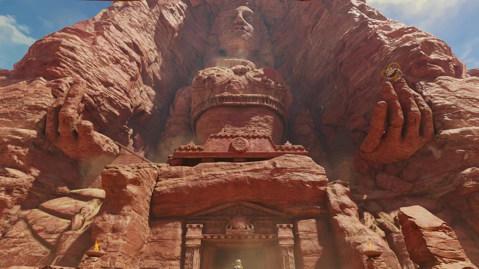 Low-angle view of the Desert Colossus: a colossal seated figure carved from red sandstone, serene face at the top, two enormous hands cupped on either side, and the ornate Spirit Temple entrance with lit torches at its base, a tiny figure standing in the doorway