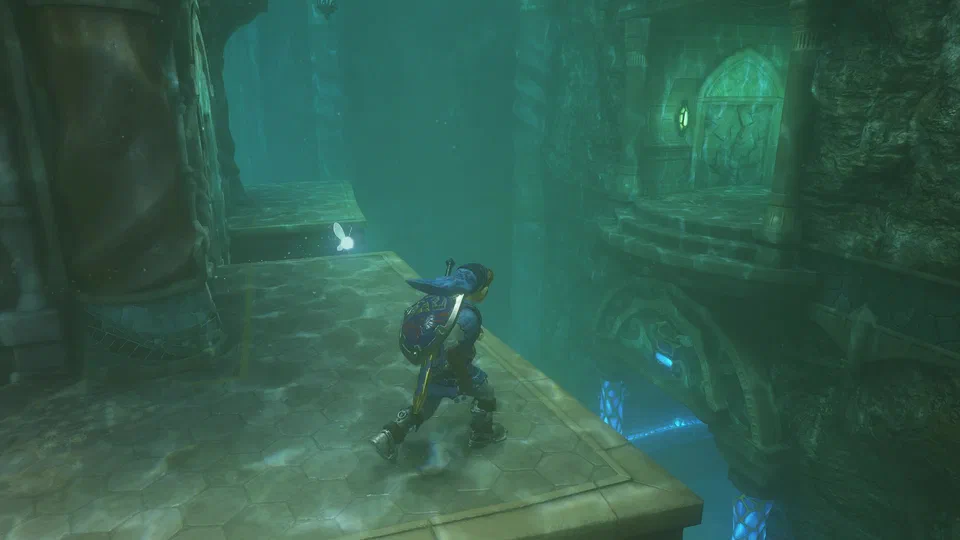 Adult Link in the blue Zora Tunic and heavy grey Iron Boots walking along a hexagonal-tiled ledge fully underwater in the flooded Water Temple, teal light caustics everywhere, carved columns and a lantern-lit doorway on a ledge across the gap
